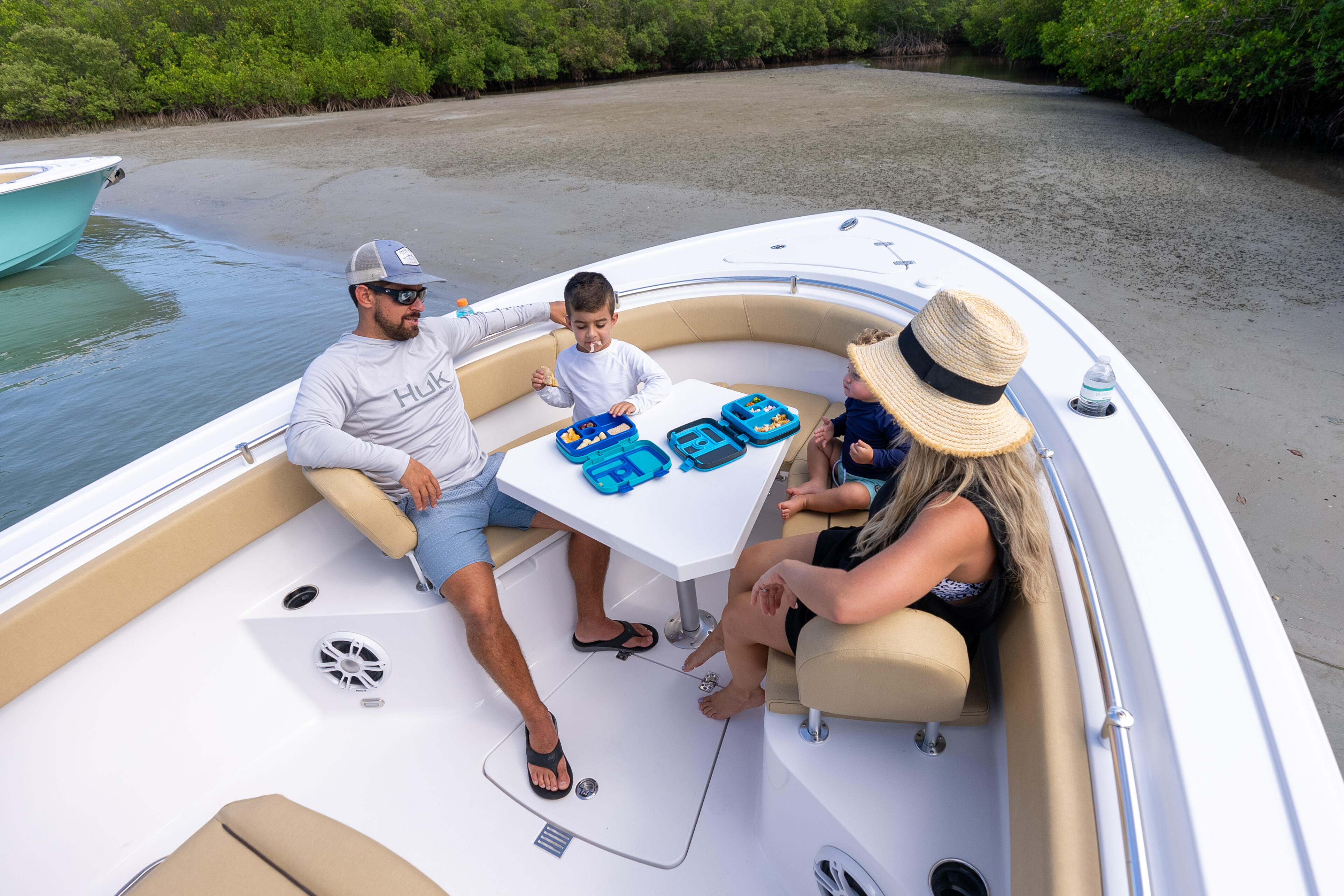 Family of four relaxing in a boat near a sandy mangrove shoreline, with blue coolers on the cabin table