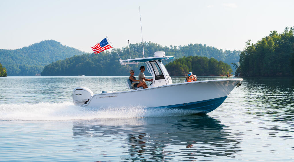 White center console boat running through clear lake waters with mountains and forested shoreline in background, American flag flying from hardtop