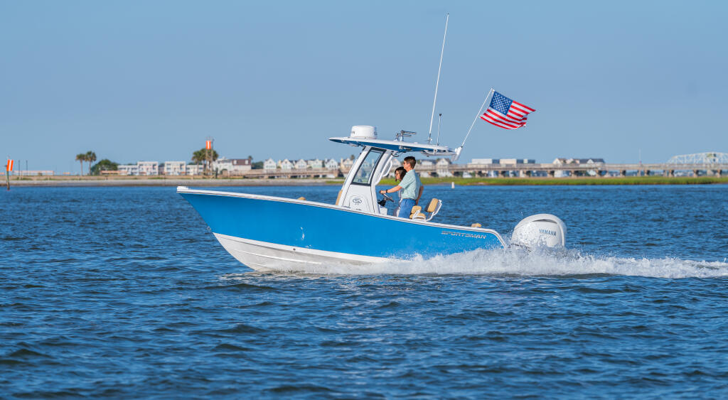 Blue and white center console boat running through calm waters with American flag and coastal homes visible in background