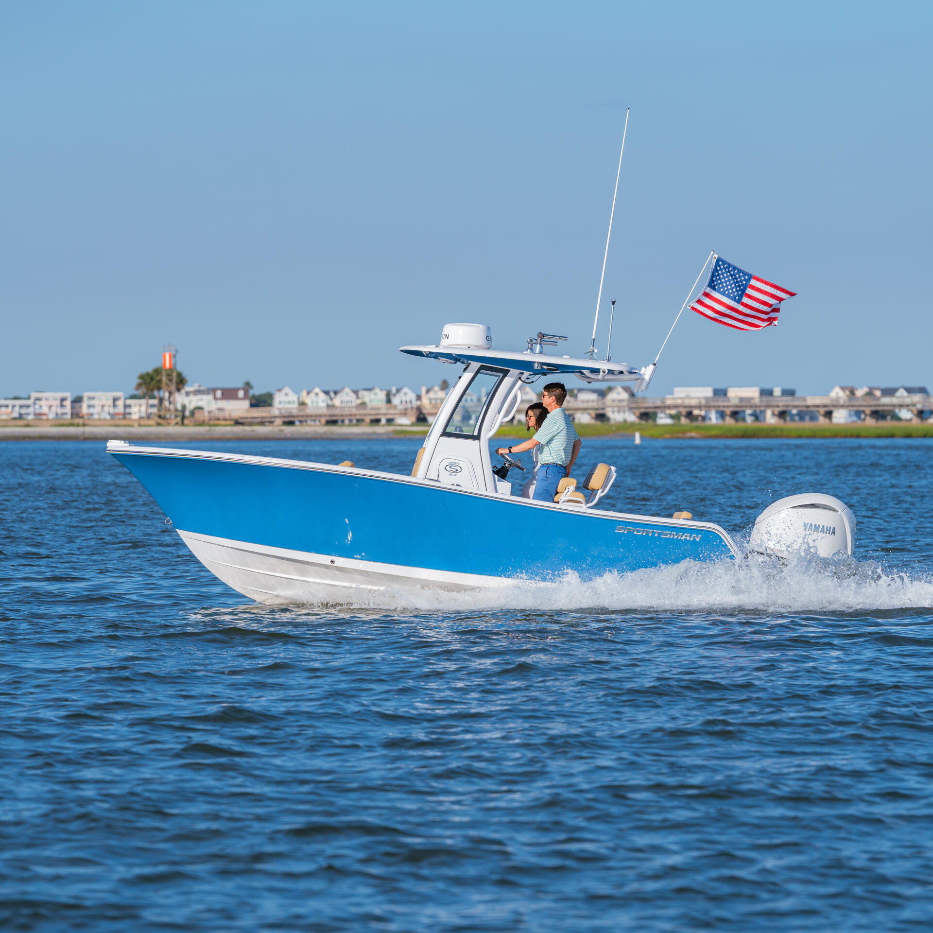 Blue and white center console boat running through calm waters with American flag and coastal homes visible in background