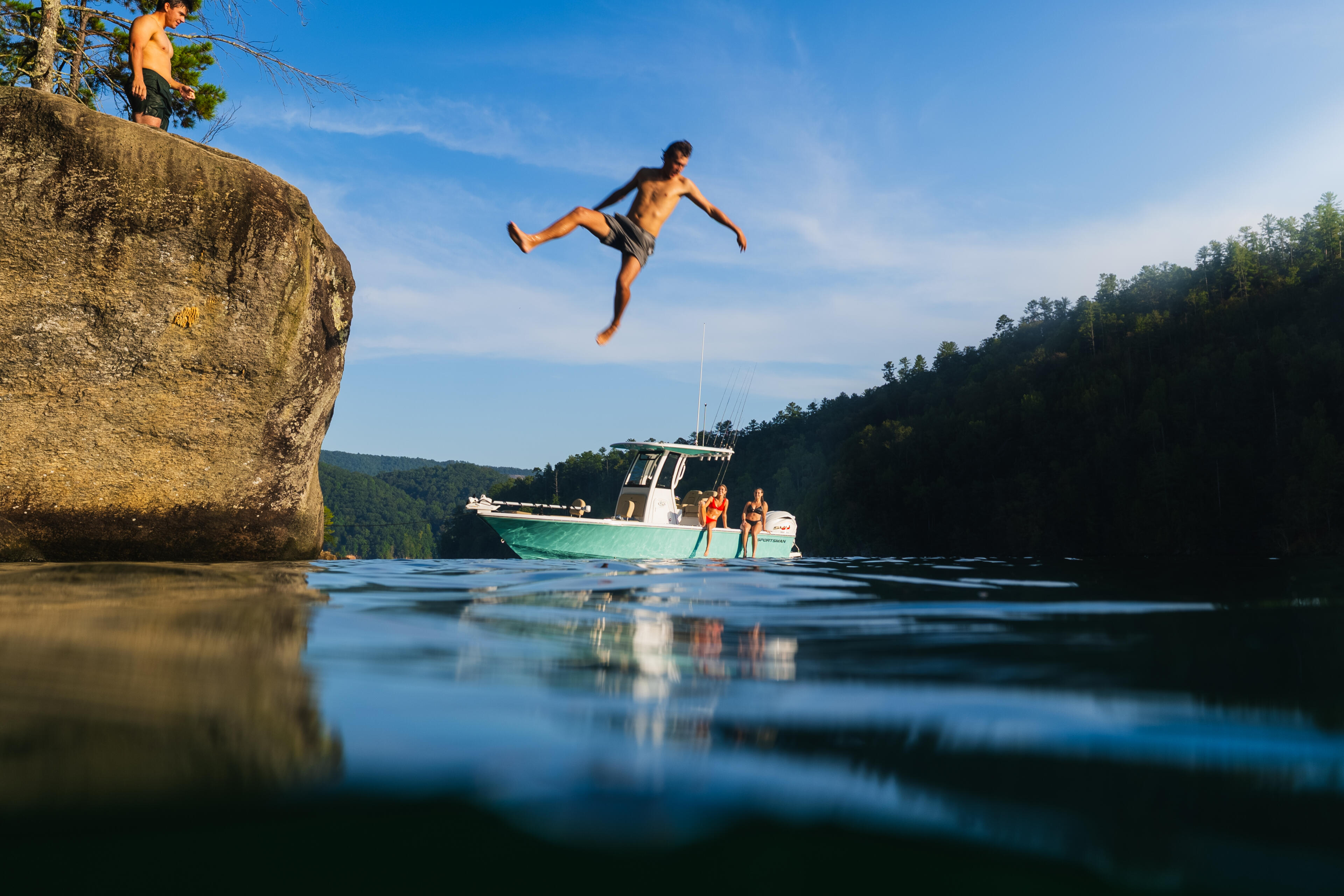 Man mid-jump from a rocky cliff toward a fishing boat anchored in clear lake water surrounded by forested hills