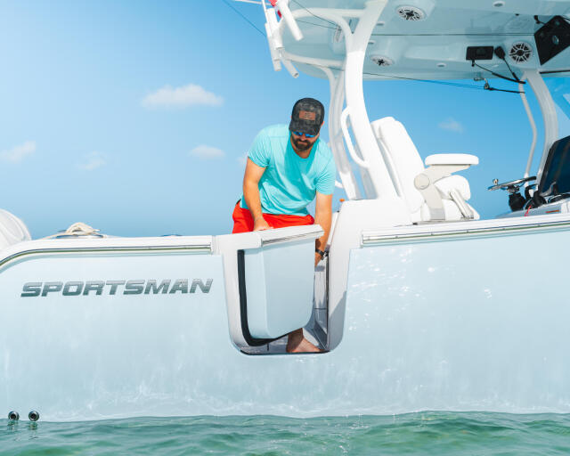 Man in turquoise shirt leaning out of Sportsman boat door at shallow sandbar with clear water and blue sky