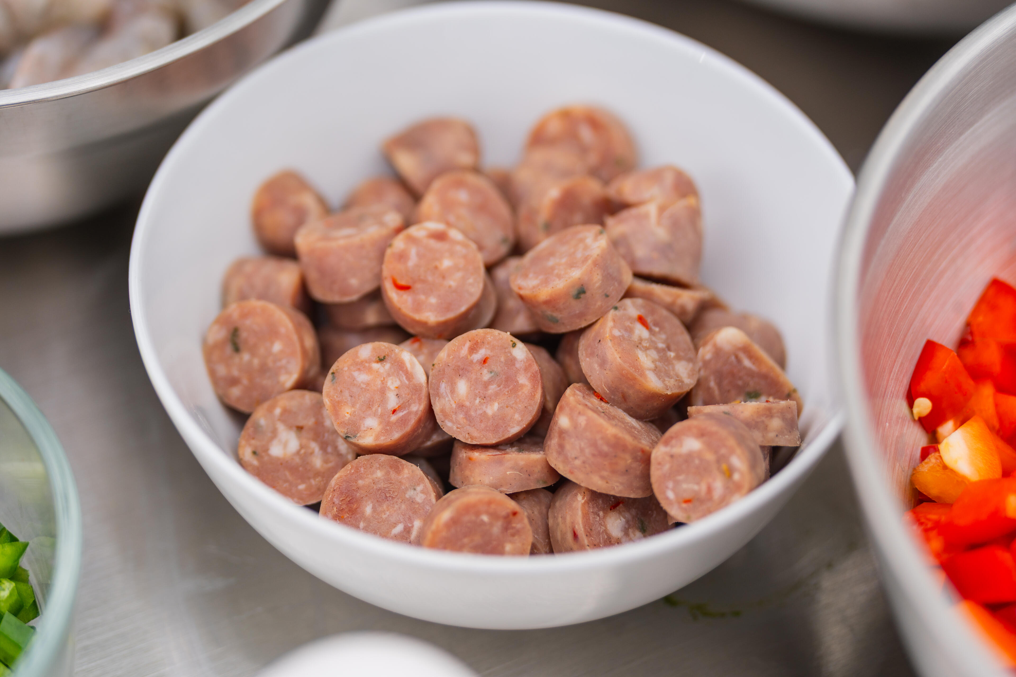 Sliced smoked sausage in a white bowl alongside fresh red bell peppers and other ingredients