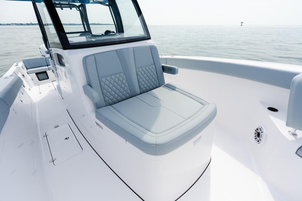 Helm seating area of a white recreational fishing boat with gray quilted upholstery and hard-top canopy overlooking calm water