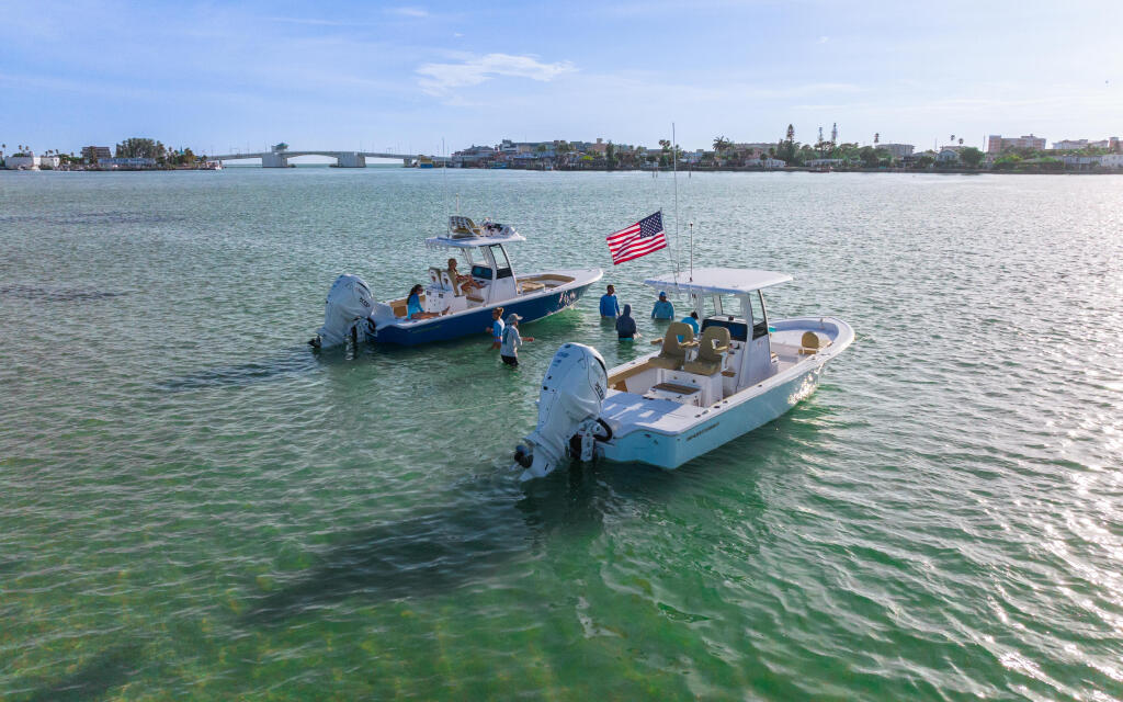 Two boats anchored in shallow turquoise water near a sandbar with families wading and enjoying the calm bay