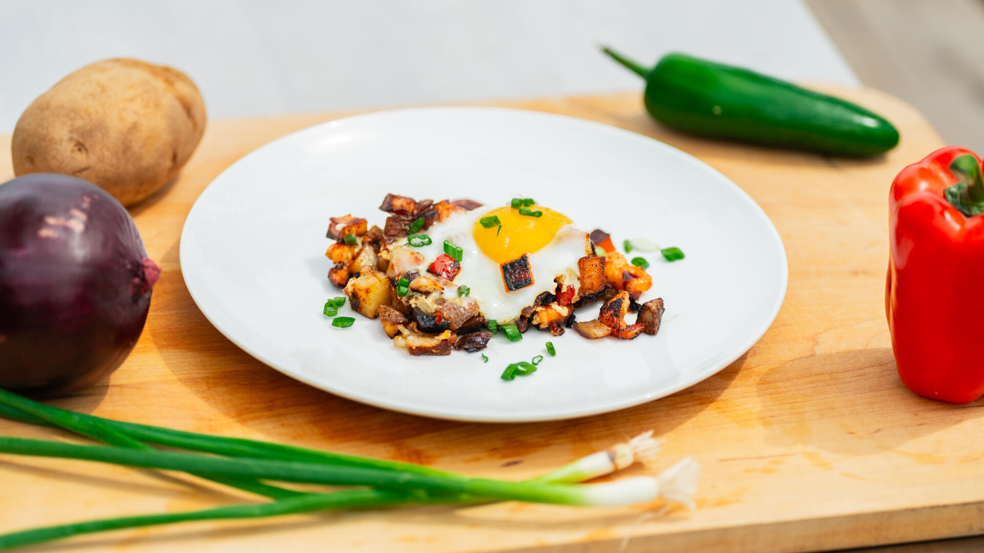 Plated lowcountry hash with fried potatoes, bacon, and a fried egg surrounded by fresh vegetables on a wooden cutting board