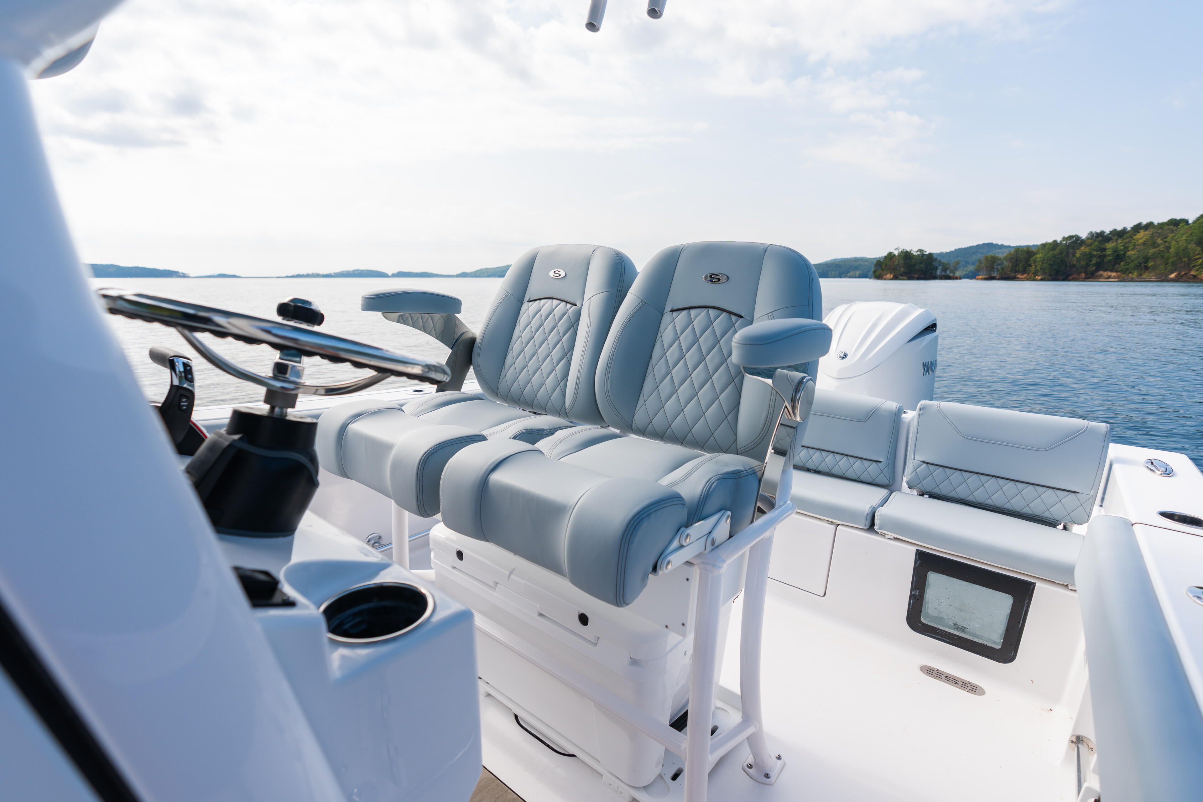 Helm area of boat with quilted captain's seats, electronics display, and calm lake with forested shoreline in background
