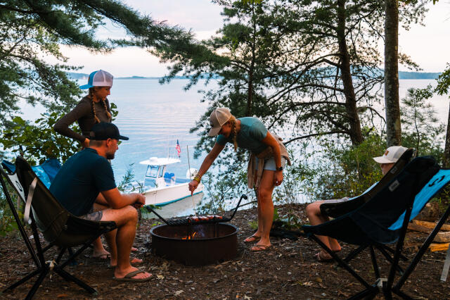 Friends gathered around a campfire by the water with a boat visible across the lake