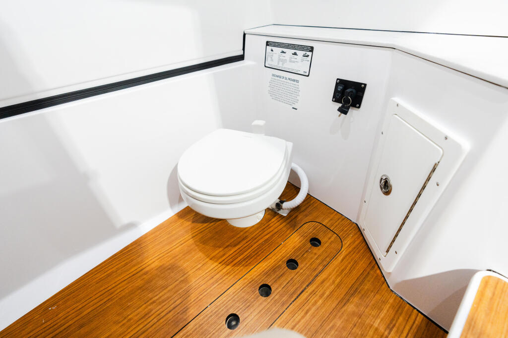 Compact boat cabin head with white toilet, wooden flooring, and white fiberglass walls with electrical panel and storage