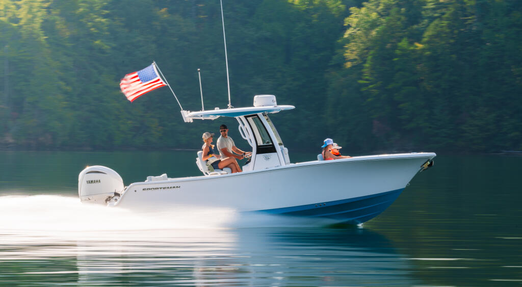 White and blue center console boat with American flag running on calm lake surrounded by forested shoreline