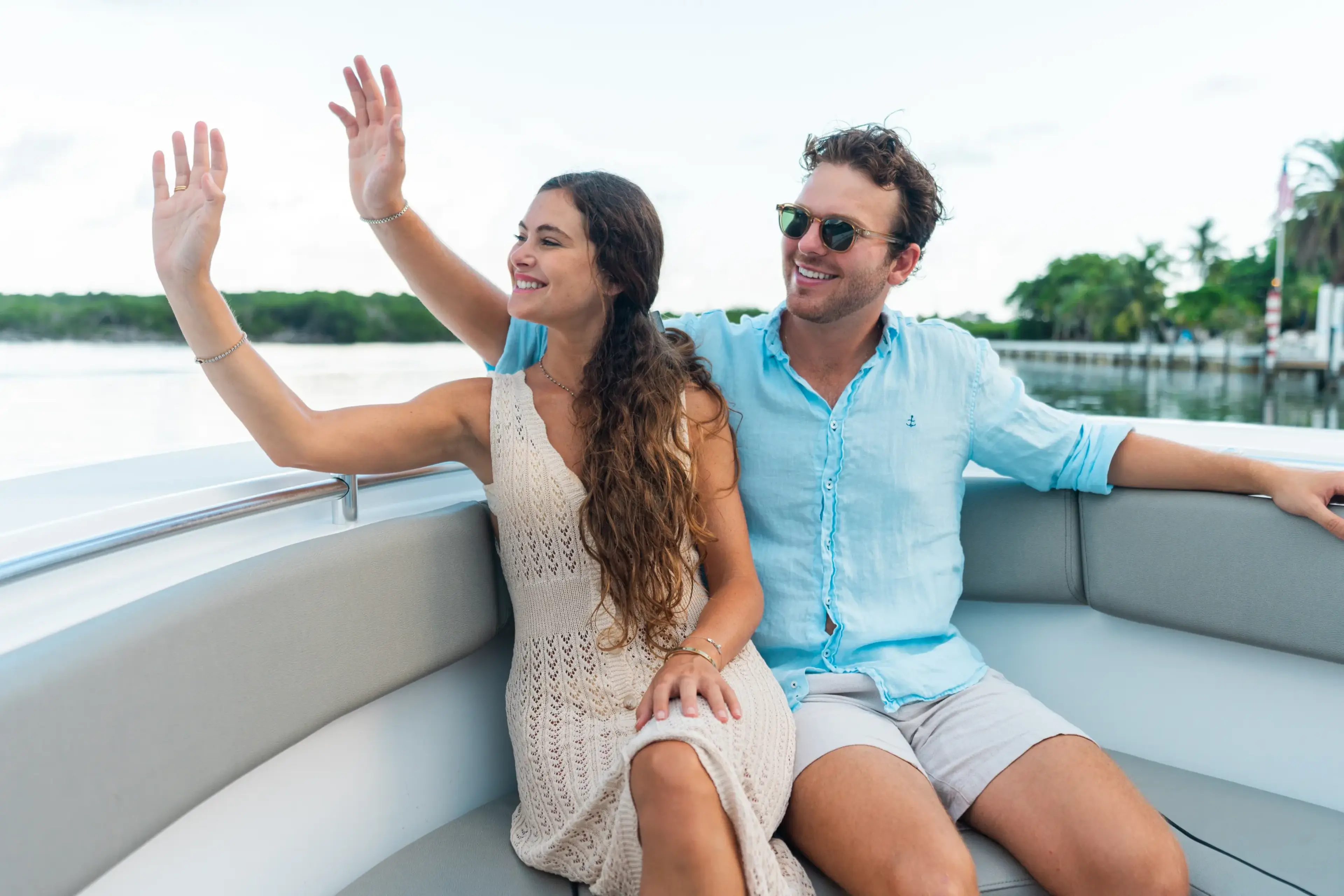 A couple on a boat waves enthusiastically while cruising past palm trees and waterfront docks