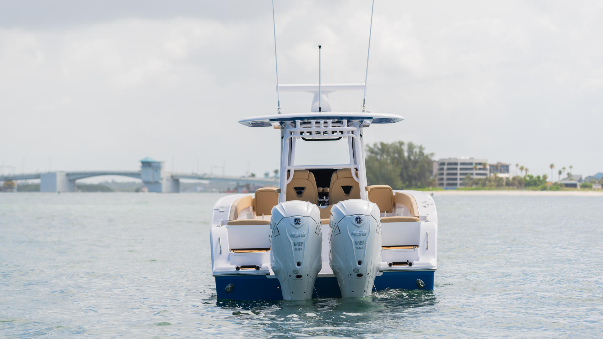 Center console boat with twin outboard engines viewed from behind, anchored in calm coastal waters near a bridge