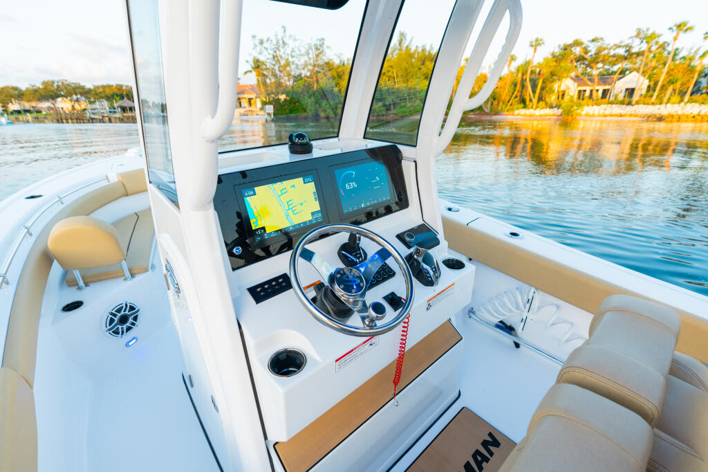 Modern boat helm with dual touchscreen displays, steering wheel, and tan seating overlooking a calm waterway